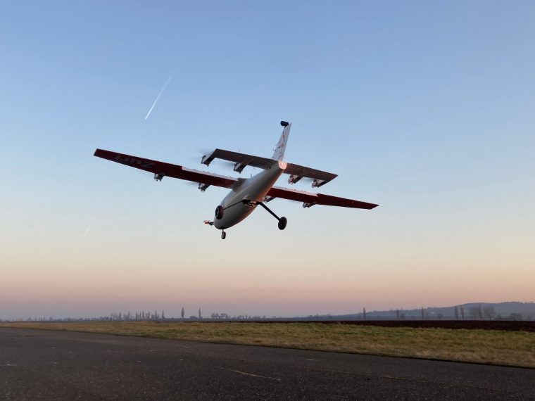 The Zuri V1NG subscale model airborne low over the airfield at dusk, landing gear down, ploughed fields and a low ridge on the horizon.